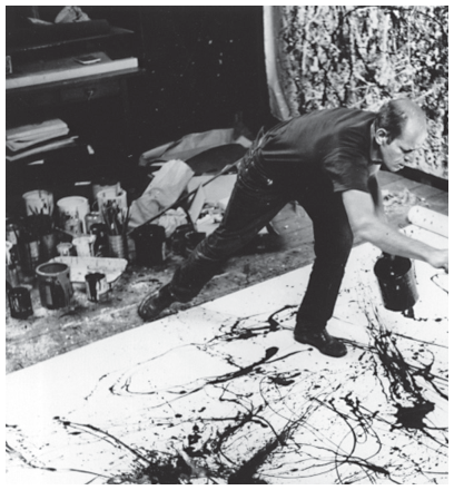 Fotografia em preto e branco. Em um ateliê, um homem se inclina sobre uma grande tela no chão. Ele estende o braço direito e segura um recipiente de tinta. A tela está coberta de respingos e fios de tinta escura. Latas e materiais de pintura aparecem à esquerda.