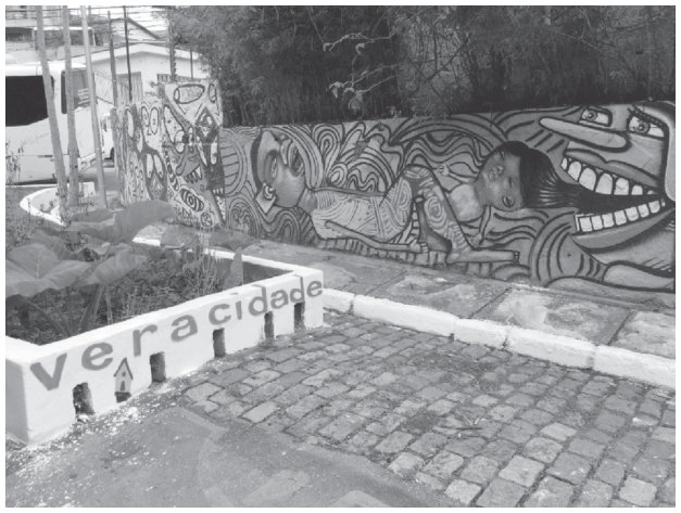 Fotografia de uma rua de paralelepípedos. Um muro à margem da rua está coberto por grafites: uma menina deitada, desenhos de linhas onduladas e um rosto estilizado com dentes à mostra. À frente, uma barreira baixa de concreto traz a inscrição “ver a cidade”.