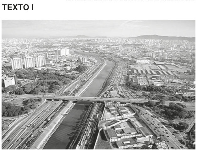 Fotografia aérea em preto e branco do Rio Tietê, em São Paulo. O rio, de margens retificadas, atravessa uma área densamente urbanizada. Há vias expressas paralelas às duas margens, uma ponte sobre o rio e muitos prédios e galpões ao redor.