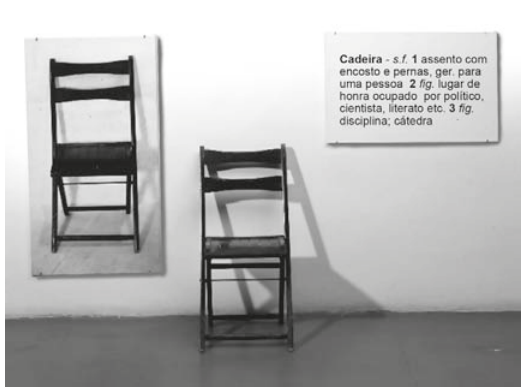 Fotografia de uma instalação com três elementos lado a lado: à esquerda, uma fotografia em preto e branco de uma cadeira; no centro, uma cadeira de madeira; à direita, um quadro com o verbete: “Cadeira — assento com encosto e pernas, geralmente para uma pessoa; lugar de honra ocupado por político, cientista ou literato; disciplina, cátedra”.