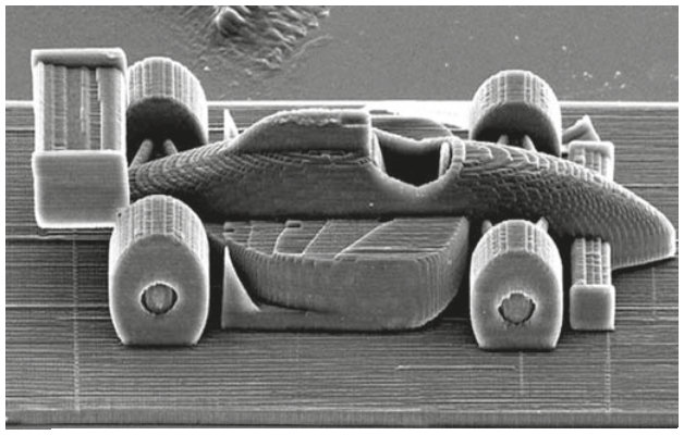 Fotografia microscópica em tons de cinza de uma miniatura de carro de Fórmula 1, vista de frente e um pouco de cima. Aparecem quatro rodas largas, carroceria aerodinâmica, assento central e aerofólio traseiro. Não há escala ou medidas na imagem.