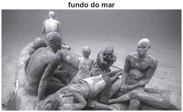 Fotografia de uma instalação no fundo do mar. Esculturas de pessoas, incluindo crianças, estão amontoadas numa balsa: algumas sentadas, outras deitadas ou debruçadas na borda. Ao redor, vê-se o fundo arenoso. Acima da fotografia, lê-se “fundo do mar”.