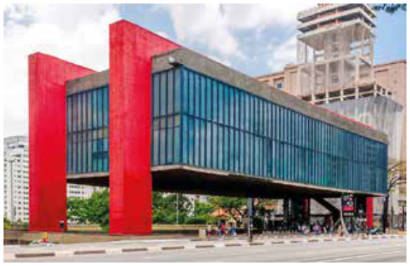 Fotografia de um edifício alongado, com fachadas de vidro azul-esverdeado e estrutura vermelha. O bloco principal é elevado, deixando um amplo vão livre sob ele, sustentado por pilares vermelhos. Há pessoas no vão e edifícios ao fundo.