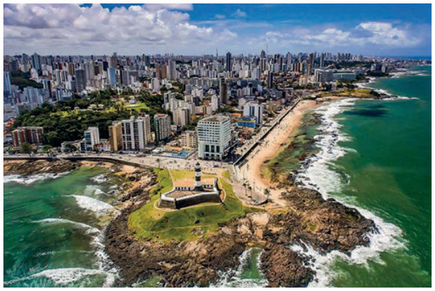 Fotografia aérea do litoral de Salvador. Em primeiro plano, um forte de alvenaria ocupa uma ponta rochosa cercada pelo mar. Ao lado, há uma praia, um calçadão e uma avenida paralela à costa. Ao fundo, numerosos edifícios de muitos andares, próximos uns dos outros, estendem-se por grande parte da área urbana.